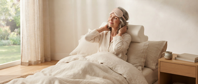 Femme allongée dans son lit, mains derrière la nuque contre un oreiller cervical, masque de sommeil sur le front